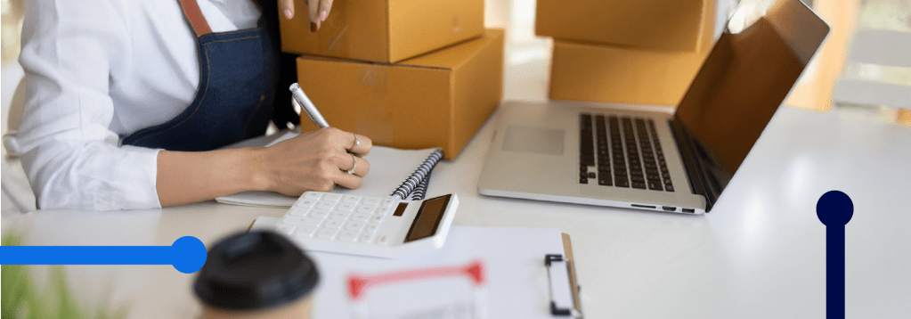 capa: DIFAL em duplicidade: É possível restituir? imagem de uma mesa cheia de caixas e um notebook. Papeis e calculadora junto com uma mão fazendo contas.